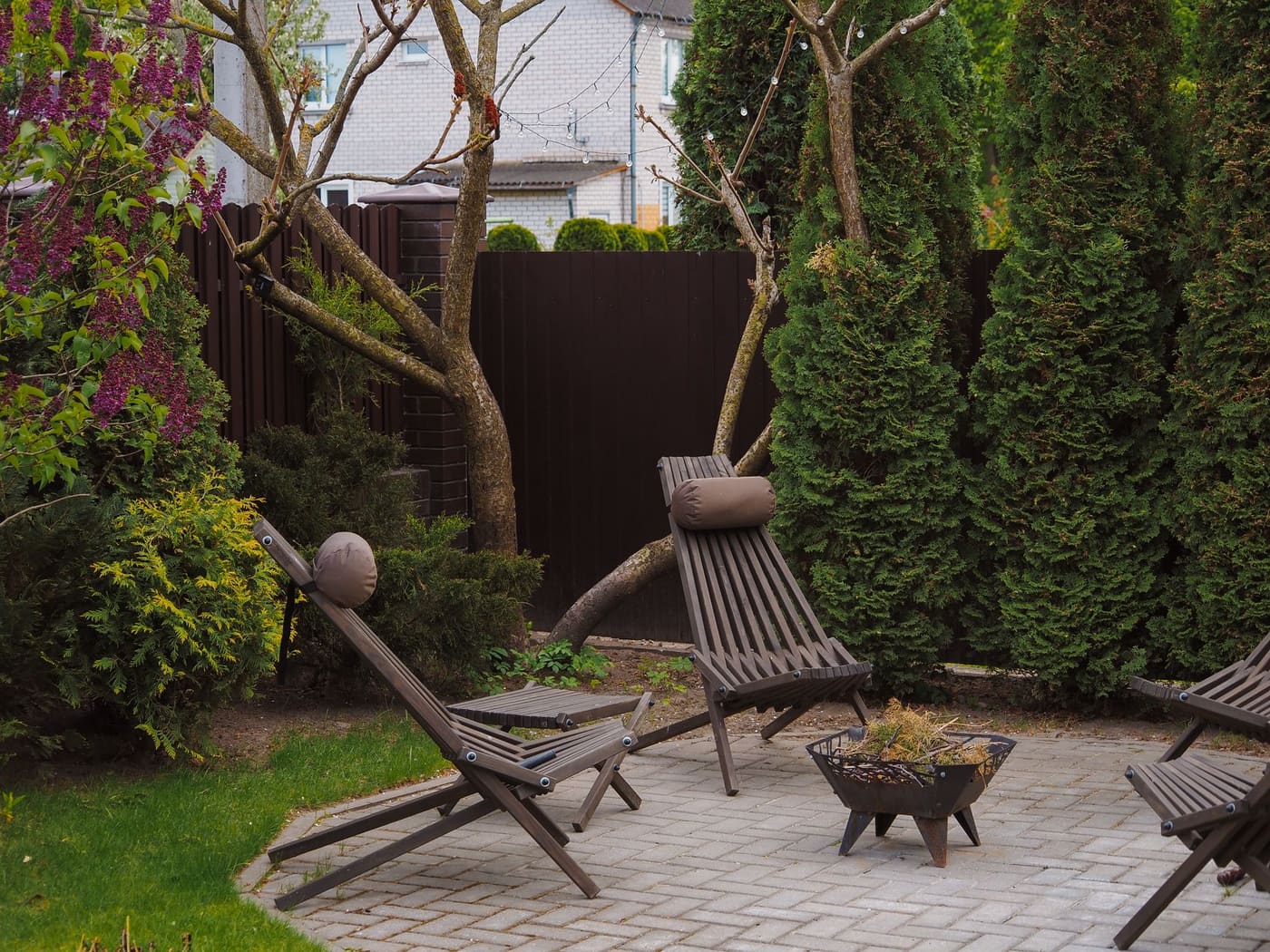 Wooden lounge chairs on a patio with tall evergreens and a dark fence behind