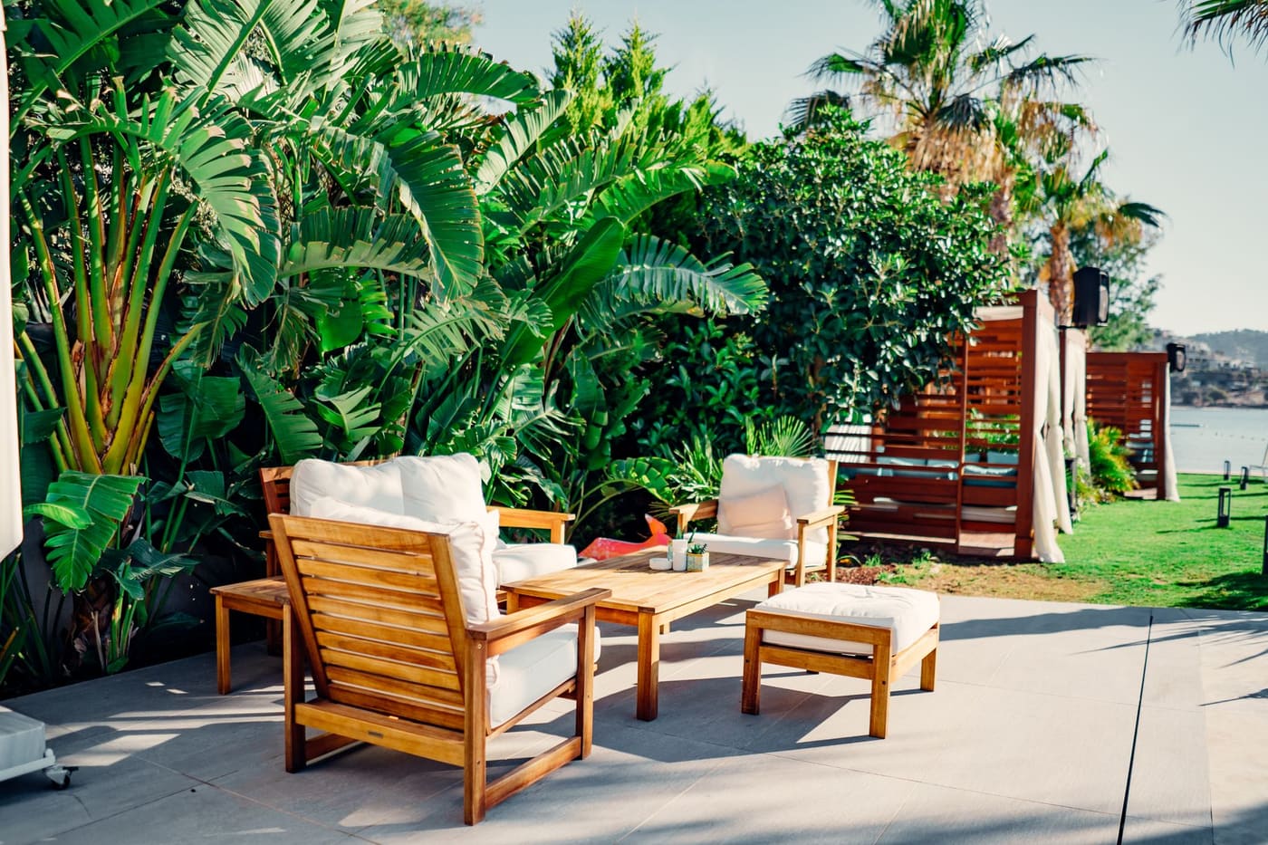 Wooden patio chairs with cushions and an ottoman beside lush green plants
